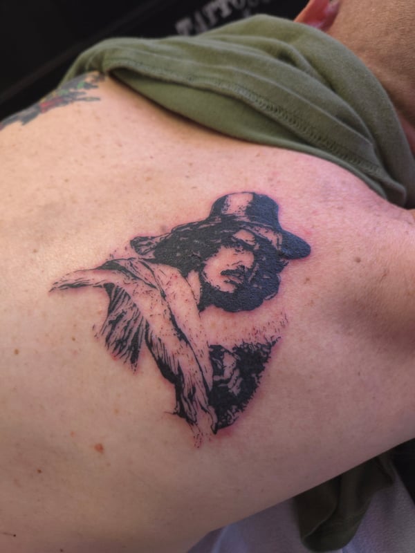 A black and grey portrait tattoo on the back, depicting a person with a hat and detailed shading — by ashynucklestattoo, los angeles