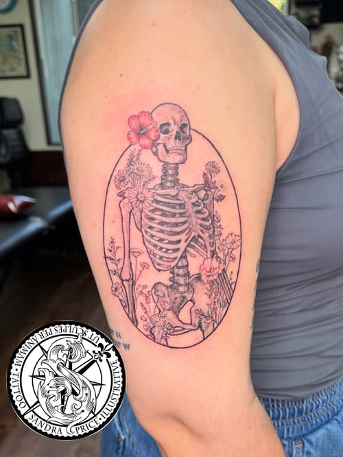 A delicate illustrative tattoo of a skeleton with flowers on the upper arm, featuring blackwork with a touch of color — by Sandra Price, los angeles
