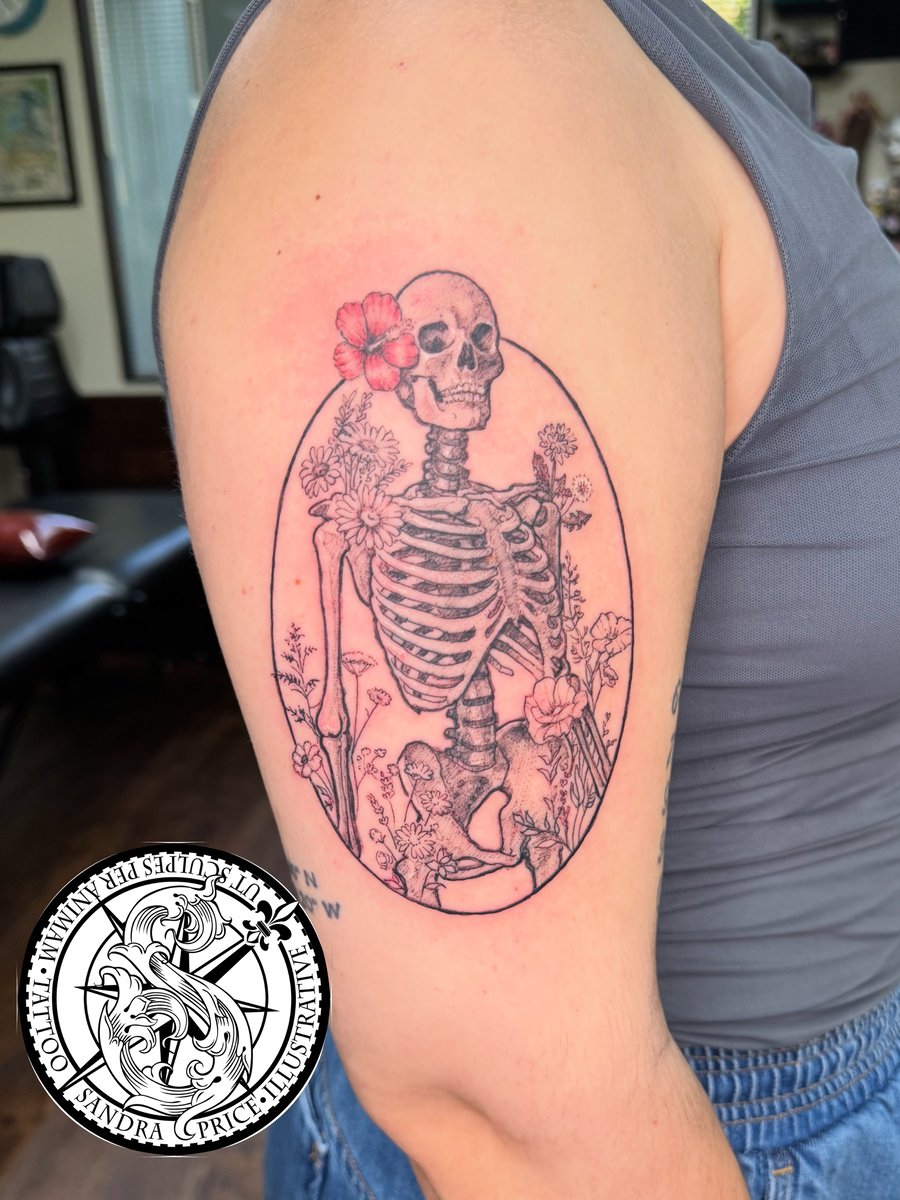 A delicate illustrative tattoo of a skeleton with flowers on the upper arm, featuring blackwork with a touch of color — by Sandra Price, los angeles