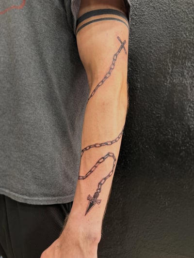 A bold blackwork tattoo of a chain with a cross and pendant on the forearm, featuring clean linework — by Kiera Garvin, los angeles