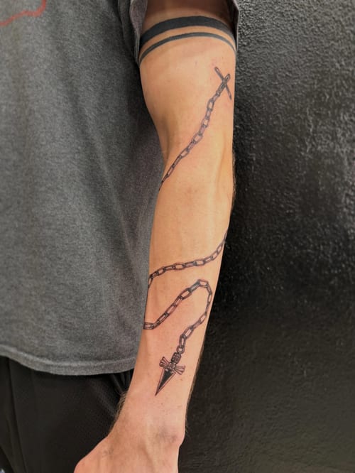 A bold blackwork tattoo of a chain with a cross and pendant on the forearm, featuring clean linework — by Kiera Garvin, los angeles