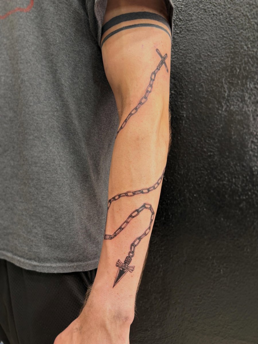 A bold blackwork tattoo of a chain with a cross and pendant on the forearm, featuring clean linework — by Kiera Garvin, los angeles