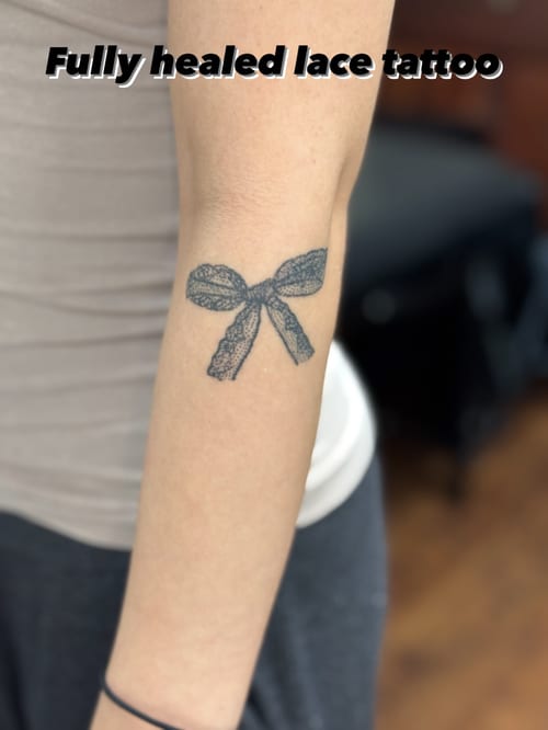 A delicate black and grey lace bow tattoo on the forearm with fine line details — by Sandra Price, los angeles