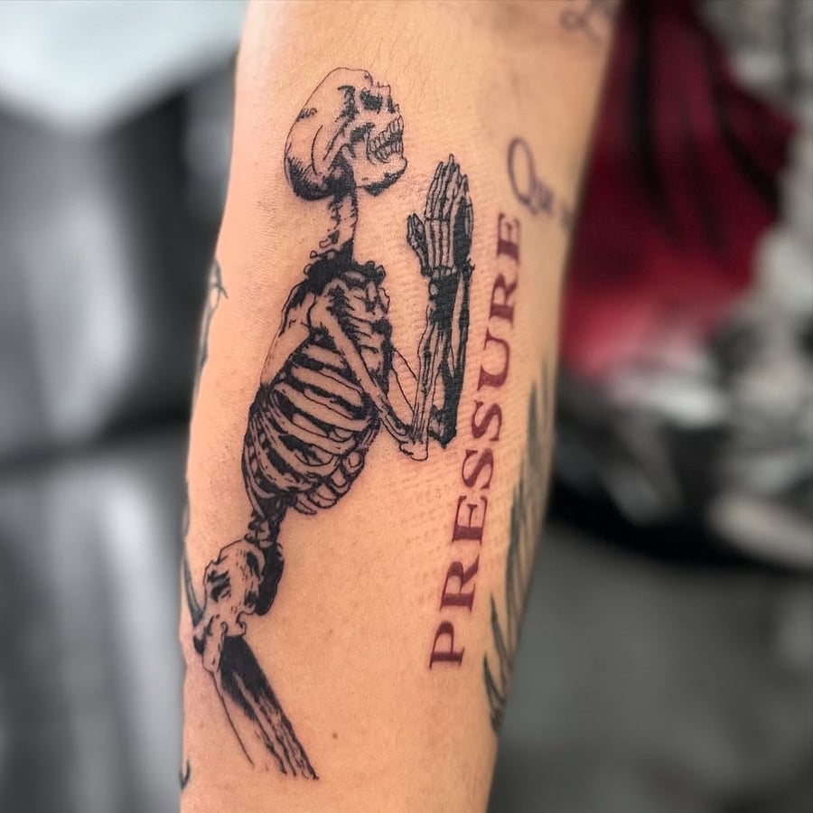 A blackwork tattoo of a praying skeleton with the word 'PRESSURE' on the arm, featuring fine line details — by Maddie Ramirez, los angeles