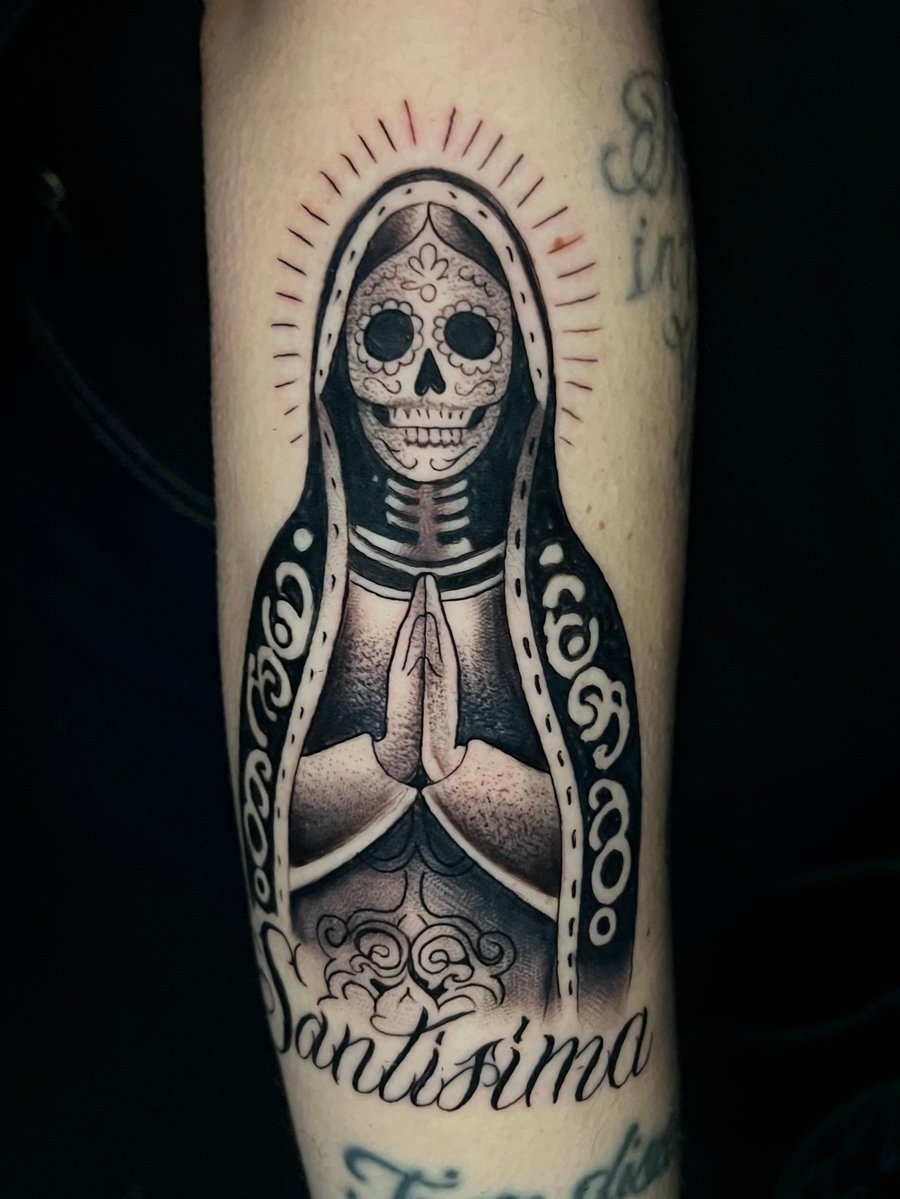 A black and grey Chicano-style tattoo on the forearm depicting a skeletal figure in a religious robe with hands in prayer, surrounded by ornamental patterns — by Josue Bravo, los angeles