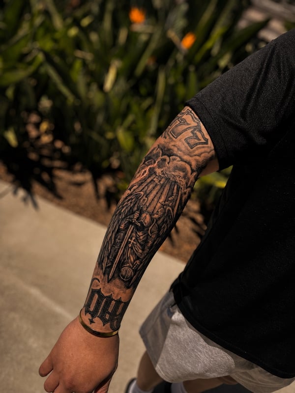 A bold black and grey tattoo of an angel with a sword on the forearm, featuring realistic shading and spiritual elements — by HEAVY INK, chino, ca