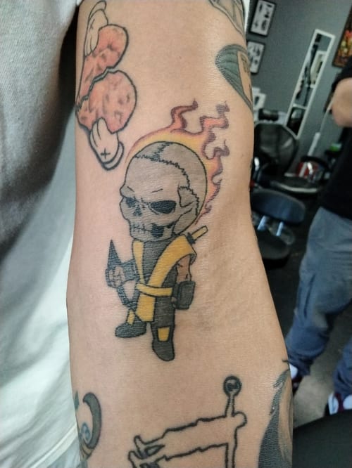 A colorful illustrative tattoo of a cartoonish skull character with flames on the arm, featuring bold outlines and vibrant colors — by anoetic.designs.ultd, los angeles