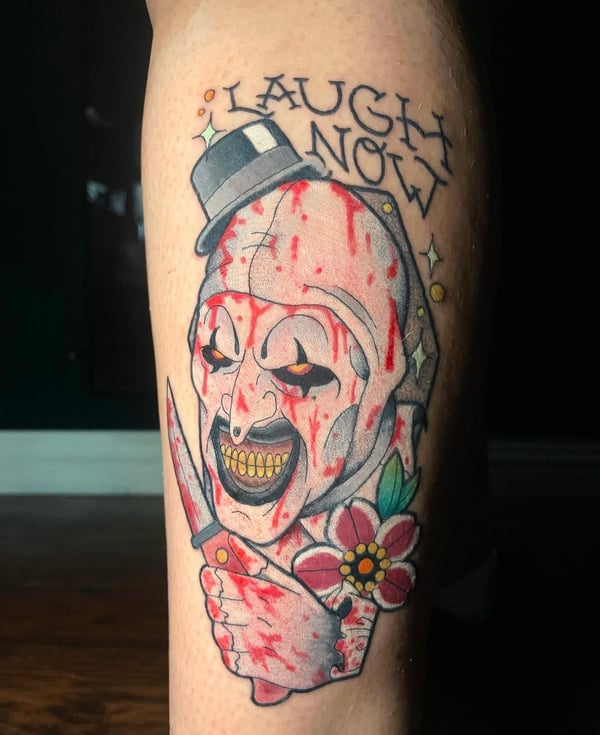 A colorful Neo-Traditional horror-themed tattoo on the leg, featuring a grinning skull with a top hat, holding a bloody dagger and a flower, with the words 'Laugh Now' above — by Jessica Linden, ri…