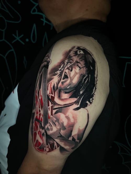 A realistic portrait tattoo on the upper arm depicting a musician playing a guitar, with a mix of black and grey shading and red accents — by Josue Bravo, los angeles