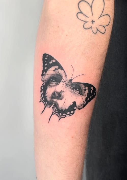 A black and grey surrealism tattoo of a butterfly with a human face on the forearm, featuring bold black lines and intricate details — by Level tattoo, los angeles