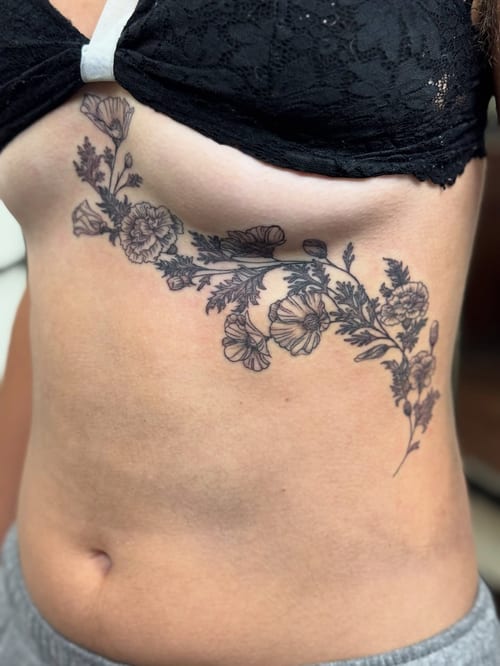 A delicate black and grey floral tattoo on the rib, featuring fine line work and intricate flower details — by Sandra Price, los angeles