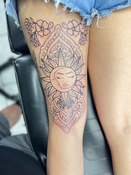 A black and grey ornamental tattoo on the thigh featuring a sun and moon motif with floral elements and intricate linework — by Mr._Folks, los angeles