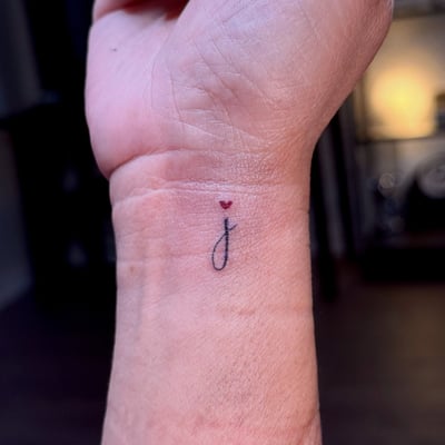 A minimalist tattoo of the letter 'j' with a small red heart above it on the wrist — by NJ TATTOOZ, los angeles