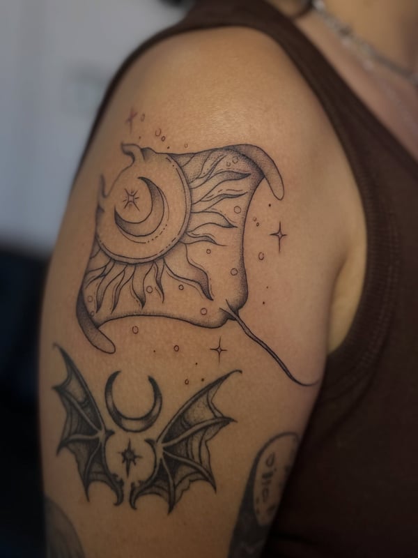 A delicate black and grey linework tattoo of a stingray with a crescent moon on the upper arm — by Maddie Ramirez, palm desert, ca