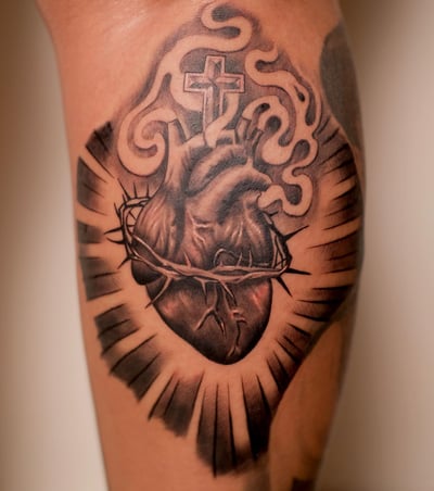 A black and grey sacred heart tattoo with a cross on the upper arm, featuring realistic shading and bold lines — by Johnny Ayala, los angeles