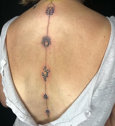 A delicate fine line tattoo along the spine featuring a lotus, sun, and moon symbols in a minimalist style — by Sandra Price, los angeles