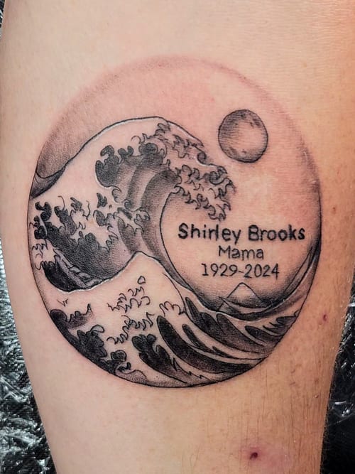 A black and grey linework tattoo on the leg depicting a wave with a memorial inscription, enclosed in a circular frame — by Dretty, los angeles