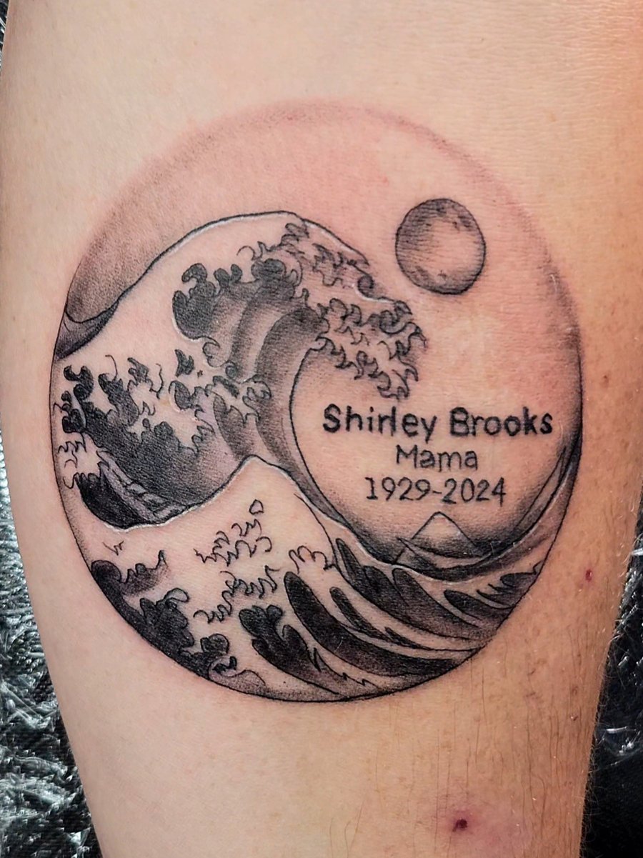 A black and grey linework tattoo on the leg depicting a wave with a memorial inscription, enclosed in a circular frame — by Dretty, los angeles