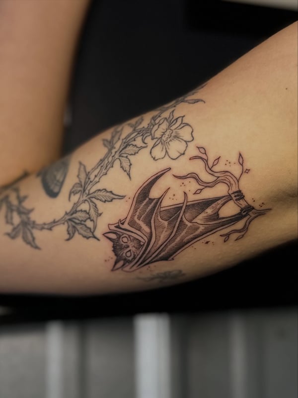 A black and grey bat tattoo with floral elements on the inner arm — by Maddie Ramirez, palm desert, ca