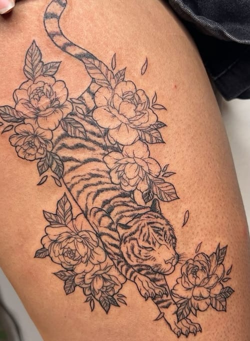 A black and grey linework tattoo of a tiger surrounded by flowers on the thigh, featuring intricate floral details and bold outlines — by NJ TATTOOZ, los angeles