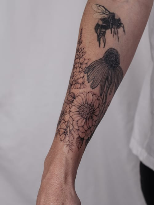 A delicate blackwork tattoo of a bee and various flowers on the forearm, featuring intricate floral details — by Melanie Benoit, los angeles