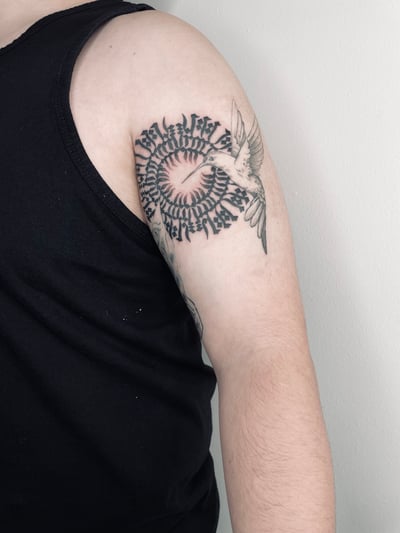 A black and grey tattoo on the upper arm featuring a bird and heart surrounded by circular lettering — by Brian Kies, los angeles