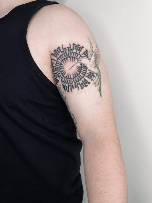 A black and grey tattoo on the upper arm featuring a bird and heart surrounded by circular lettering — by Brian Kies, los angeles