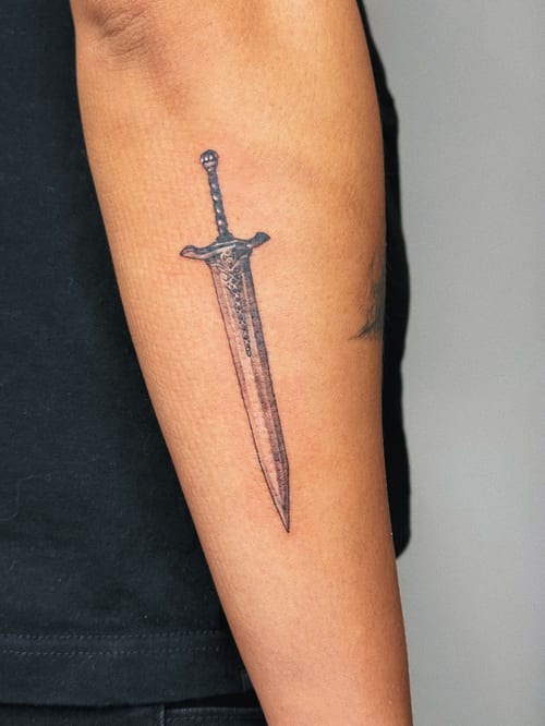 A minimalist black and grey sword tattoo on the forearm with clean lines and simple design — by lunayflorestattoo, los angeles