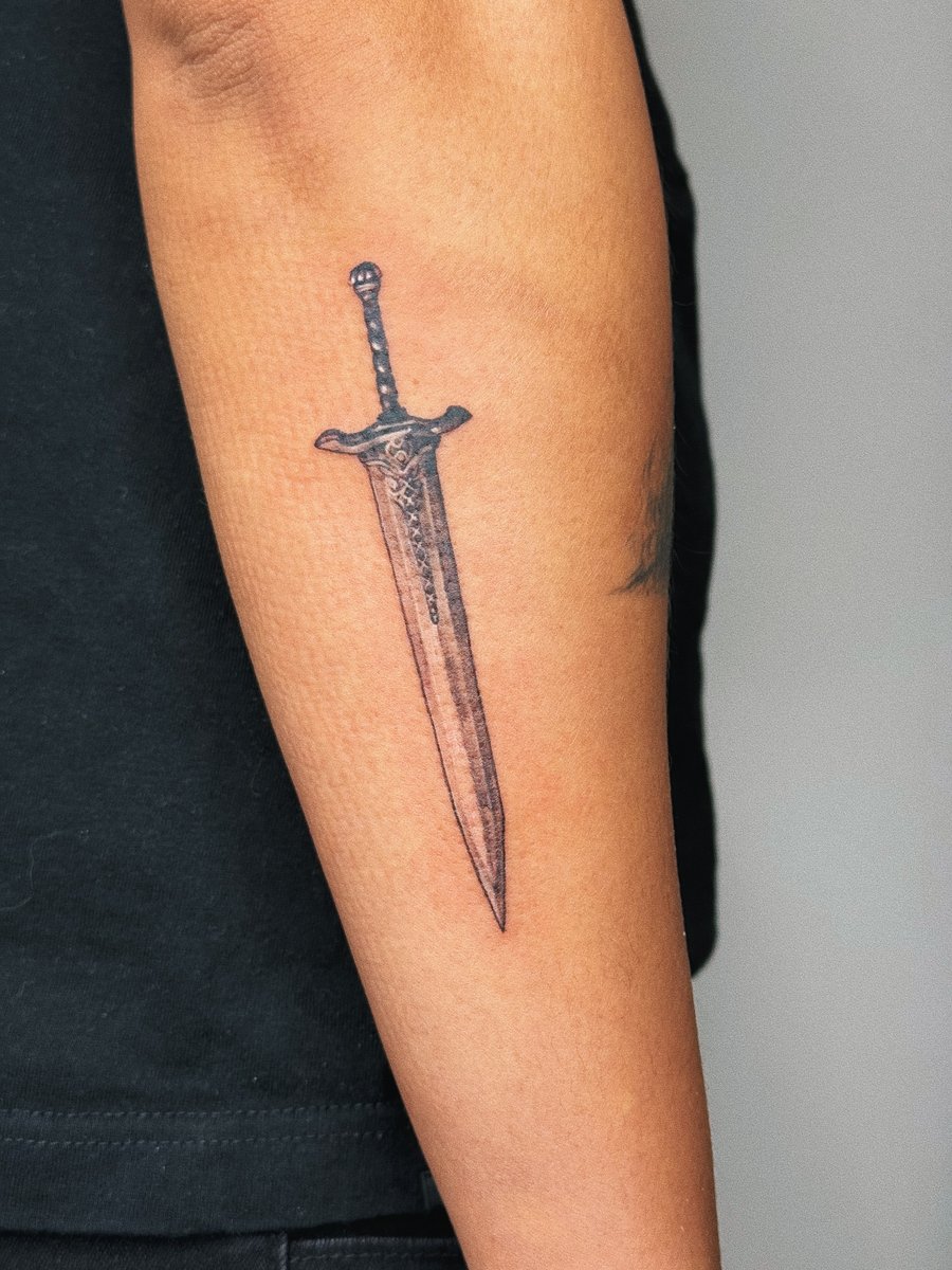 A minimalist black and grey sword tattoo on the forearm with clean lines and simple design — by lunayflorestattoo, los angeles