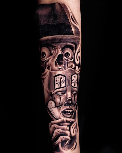 A dark black and grey Chicano style tattoo on the forearm featuring a skull and face with intricate details — by Johnny Ayala, los angeles