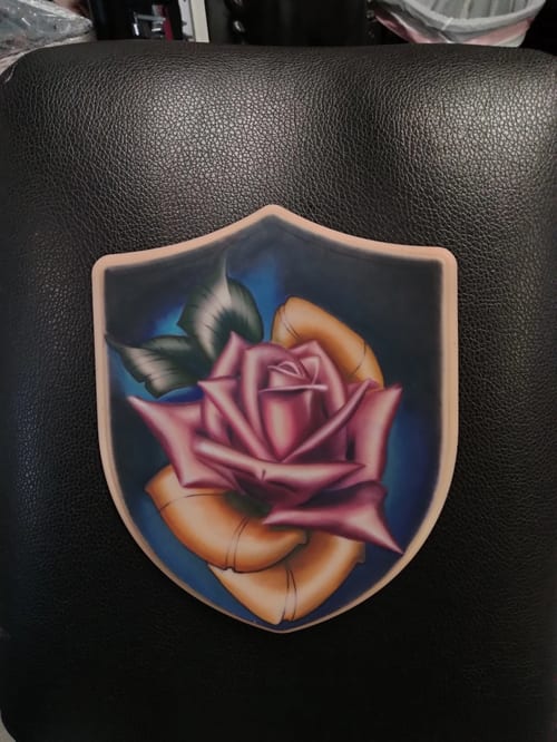 A vibrant Neo-Traditional style tattoo of a pink rose with green leaves and a yellow ribbon, featuring bold outlines and rich colors — by anoetic.designs.ultd, los angeles