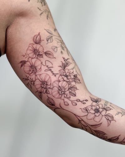 A delicate black and grey floral arrangement with script on the inner arm, featuring fine line details and a romantic feel — by Melanie Benoit, los angeles