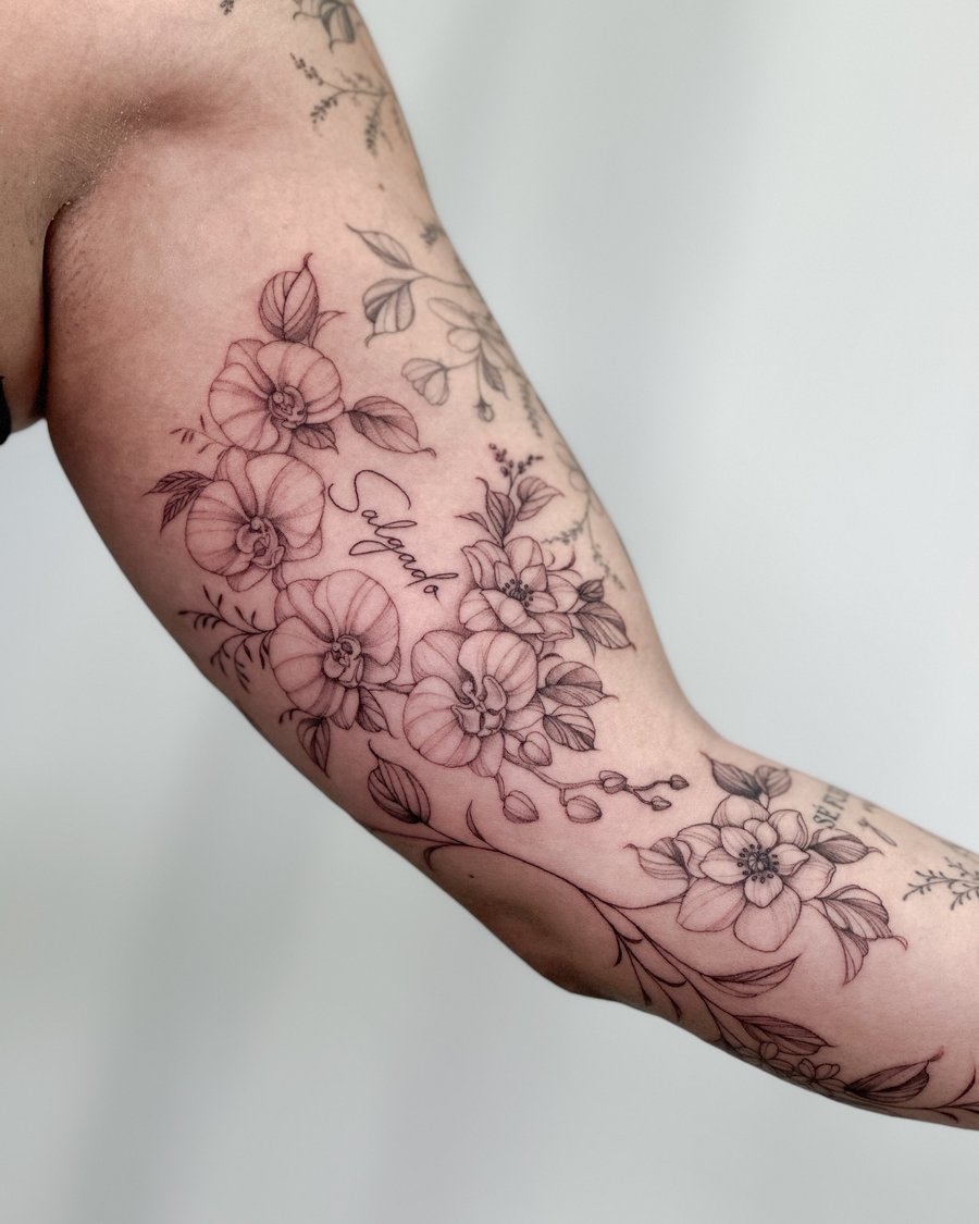 A delicate black and grey floral arrangement with script on the inner arm, featuring fine line details and a romantic feel — by Melanie Benoit, los angeles