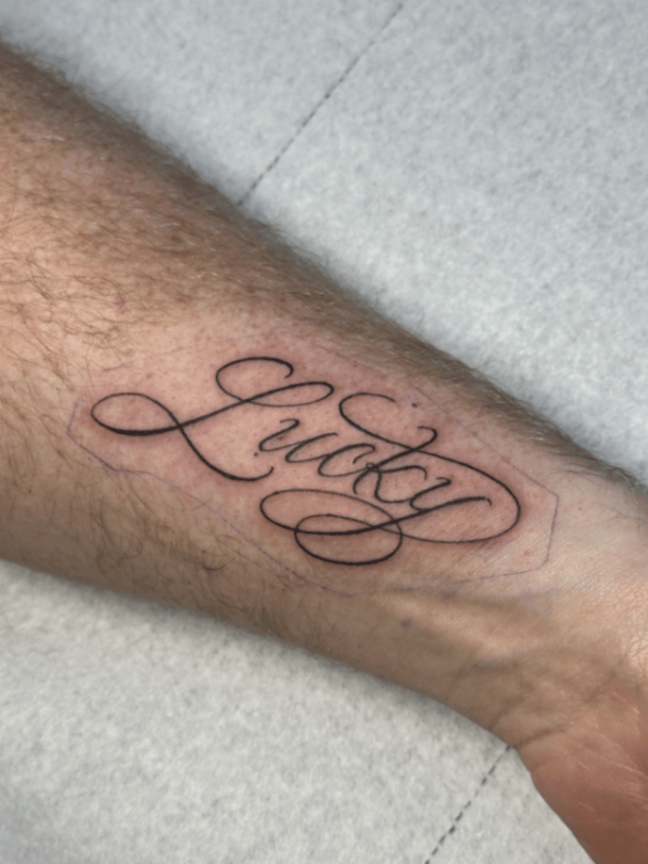 A black and grey script tattoo of the word 'Lucky' on the forearm, featuring elegant cursive lettering — by Garrick {Gman} lee, los angeles
