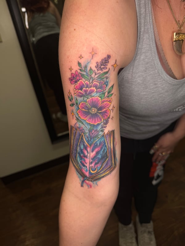 A colorful watercolor tattoo of flowers emerging from an open book on the upper arm, with a whimsical and delicate style — by Sandra Price, ventura, ca