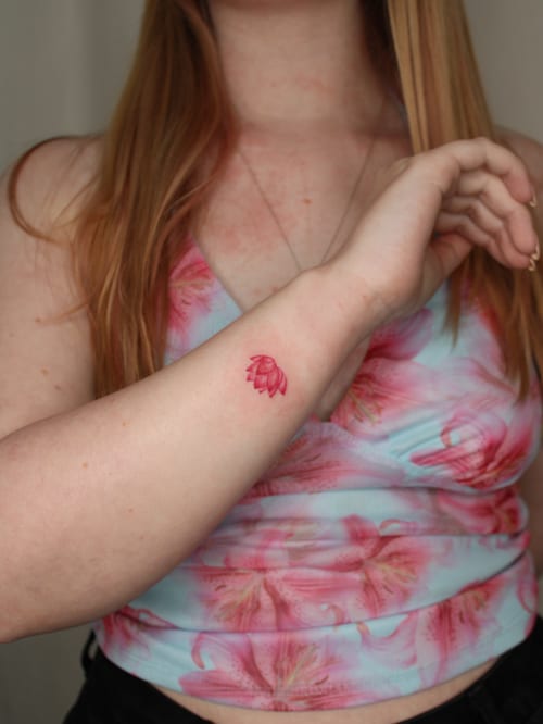 A small and delicate pink flower tattoo on the forearm, done in a minimalist fine line style — by Melanie Benoit, los angeles