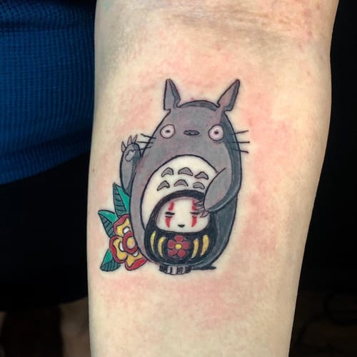 A colorful tattoo of Totoro with a daruma doll and flower on the forearm, featuring a whimsical anime style — by Sandra Price, los angeles