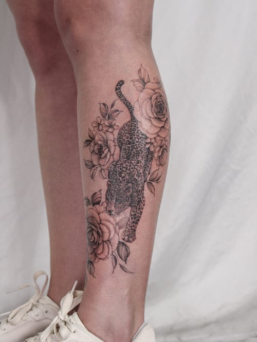 A realistic black and grey tattoo of a leopard surrounded by roses on the calf, combining animal and floral elements — by Melanie Benoit, los angeles