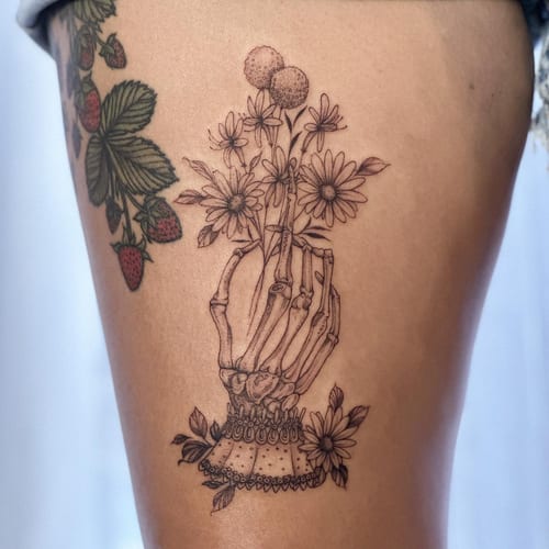 A delicate black and grey tattoo of a skeleton hand holding flowers on the thigh, featuring fine line and blackwork styles — by Melanie Benoit, los angeles