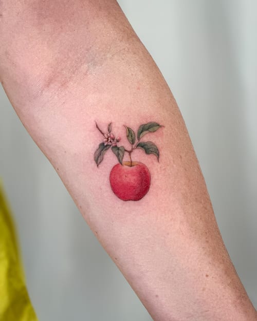 A delicate color tattoo of an apple with leaves on the forearm, rendered in a realistic style with fine line details — by Melanie Benoit, los angeles