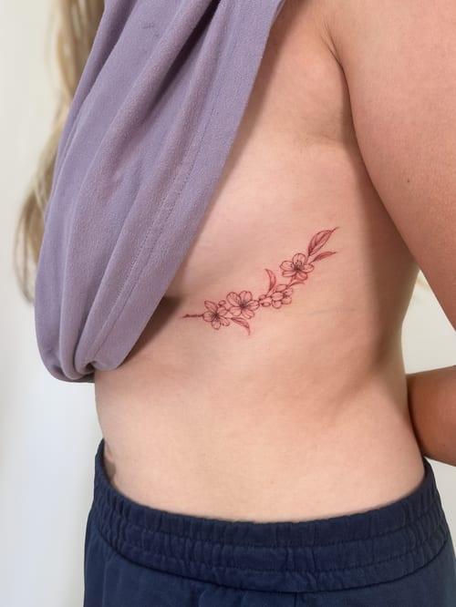 A delicate fine line floral branch tattoo in red ink on the rib, featuring minimalistic flower designs — by Melanie Benoit, los angeles