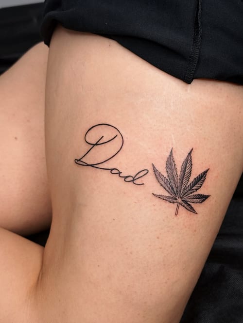 A minimalist black and grey tattoo on the thigh featuring the word 'Dad' in script alongside a leaf design — by lunayflorestattoo, los angeles
