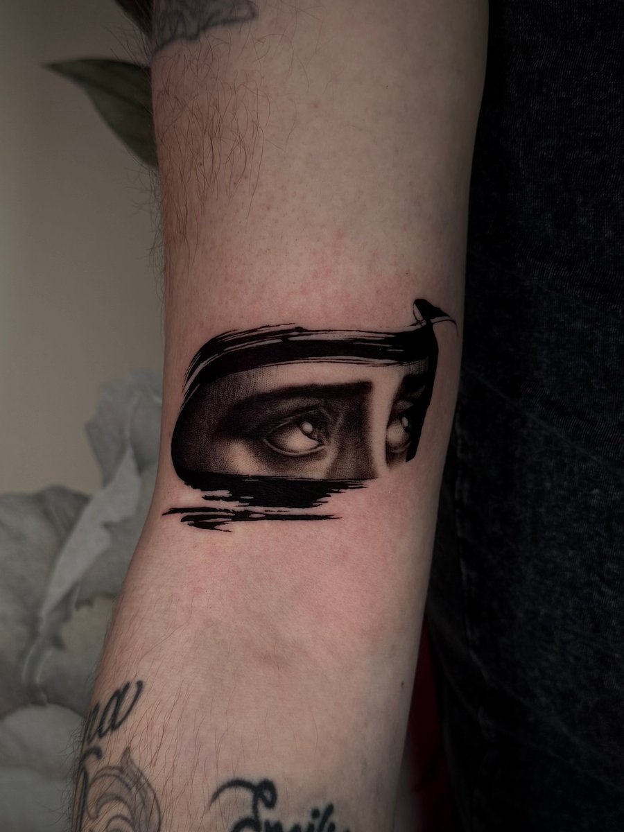 A black and grey realism tattoo of a pair of eyes with a blackwork brushstroke effect on the inner arm — by emily, los angeles