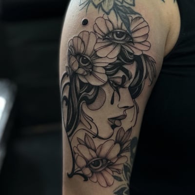 A delicate black and grey tattoo of a woman's profile with flowers and eyes on the upper arm — by Maddie Ramirez, los angeles