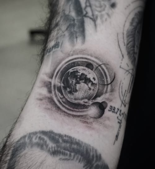A black and grey surrealistic tattoo of celestial bodies, including a detailed moon and planets, on the forearm — by Jian Liang, los angeles