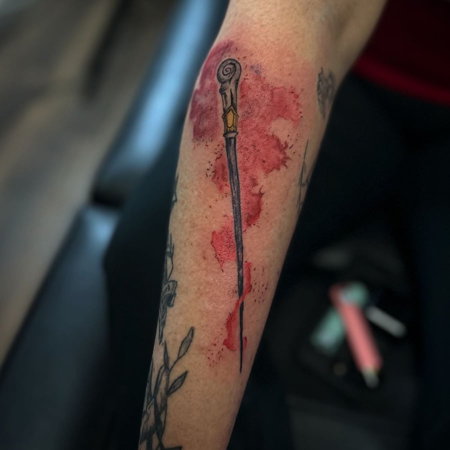 A bold dagger tattoo on the forearm with a watercolor background in mixed colors — by Maddie Ramirez, los angeles