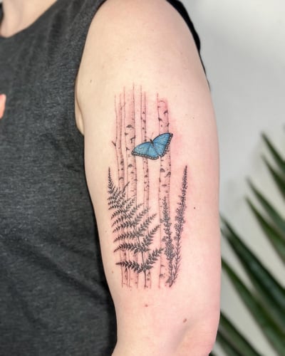 A delicate tattoo of a blue butterfly with ferns and birch trees on the upper arm, done in fine line style with mixed color and black ink — by Melanie Benoit, los angeles