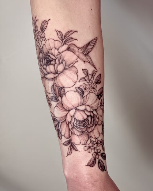 A delicate black and grey tattoo of a hummingbird and flowers on the forearm, done in a fine line style — by Melanie Benoit, los angeles