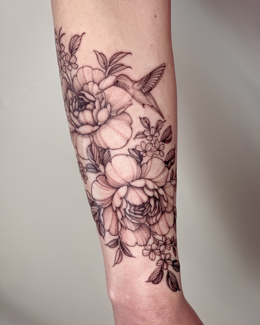 A delicate black and grey tattoo of a hummingbird and flowers on the forearm, done in a fine line style — by Melanie Benoit, los angeles