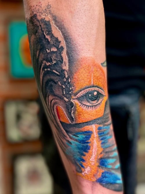 A colorful surrealistic tattoo on the forearm featuring an eye integrated into a wave with vibrant orange and blue hues — by Josue Bravo, los angeles